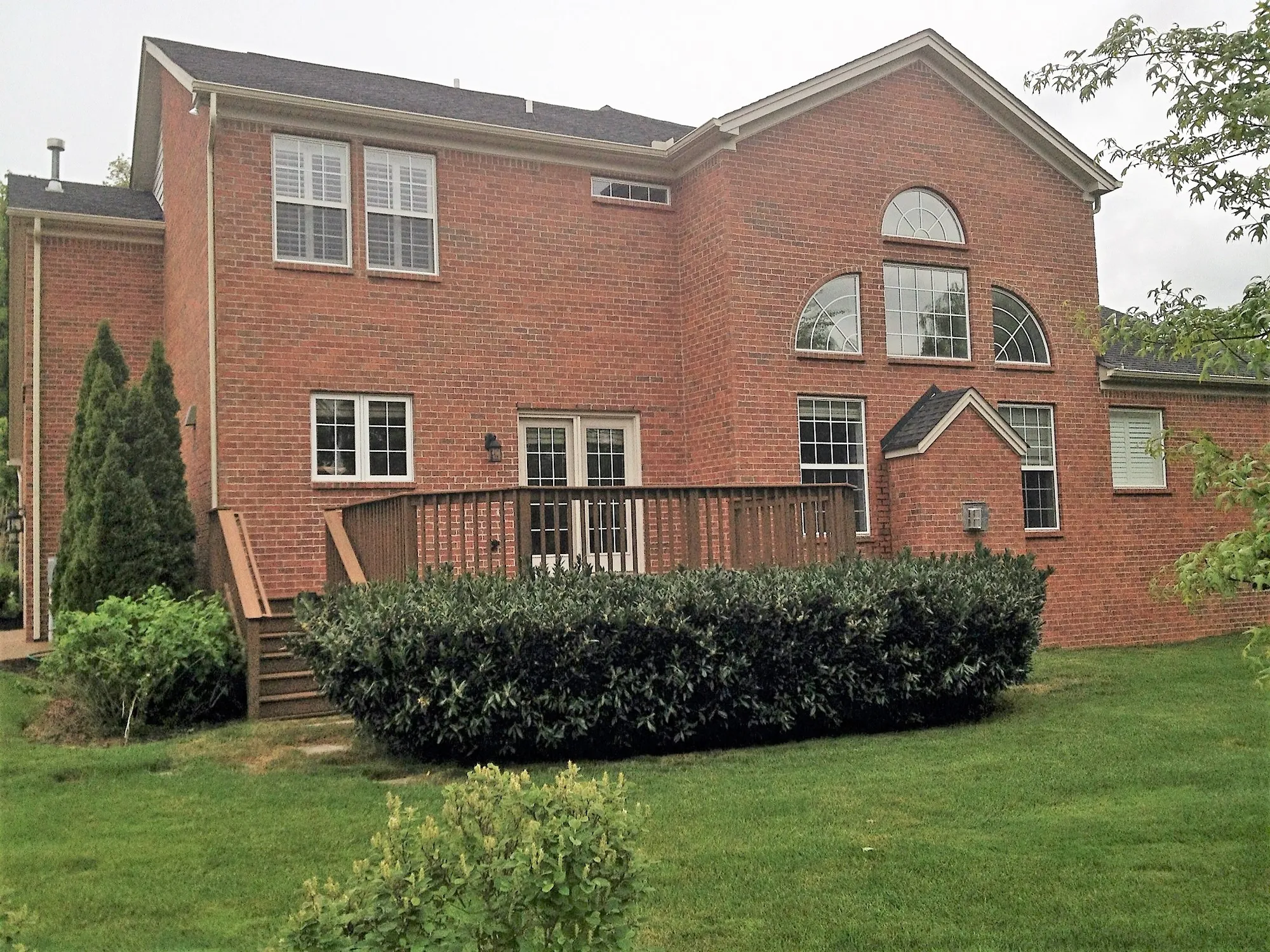 Back of a two-story red brick house with a low wood deck and shrubs