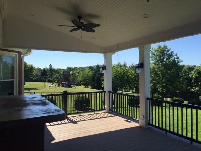Covered porch with a beadboard ceiling, a fan and black railing looking out on a lawn and trees