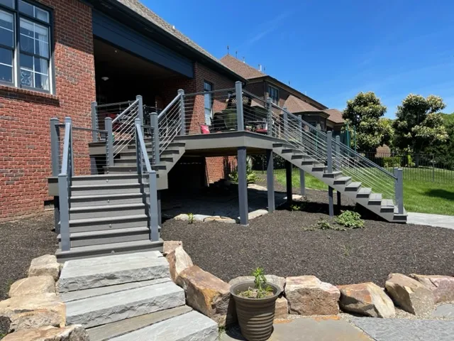 Long gray deck stairs with cable railing coming down from a brick house onto a stone landing in a mulched bed