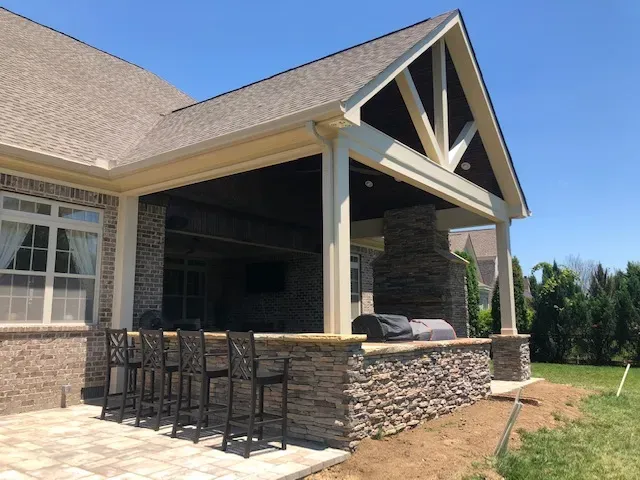 Stone outdoor kitchen bar with stools under an open gable roof attached to a brick house
