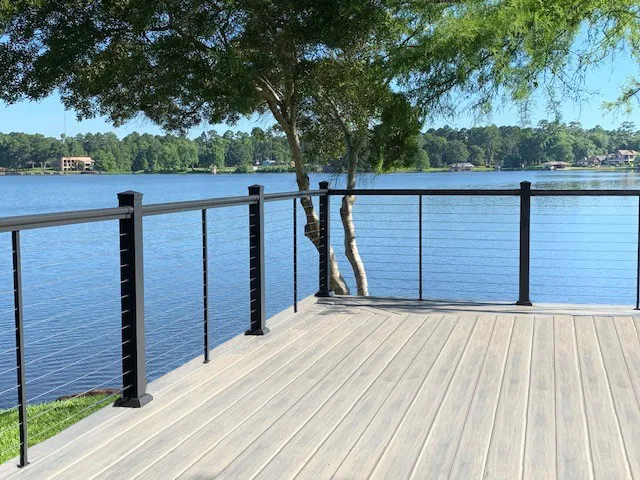 Light gray composite deck with black posts and cable railing on the edge of a lake under a tree