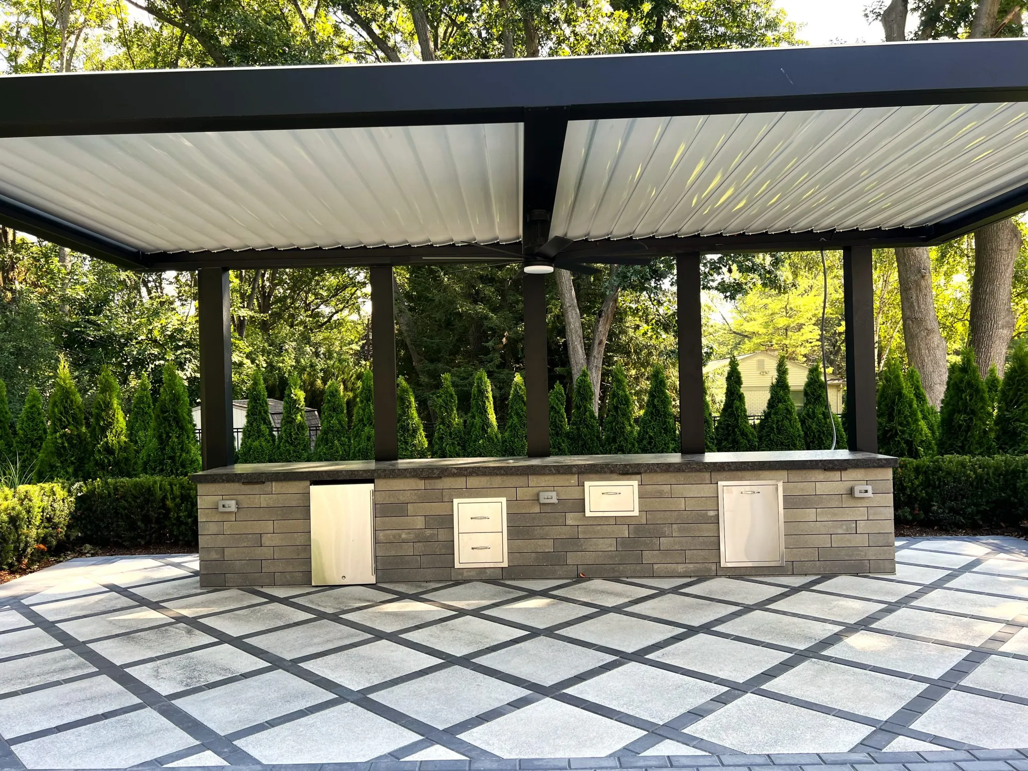 White louvered roof on dark posts over a stone outdoor kitchen on a large paver patio with evergreens behind