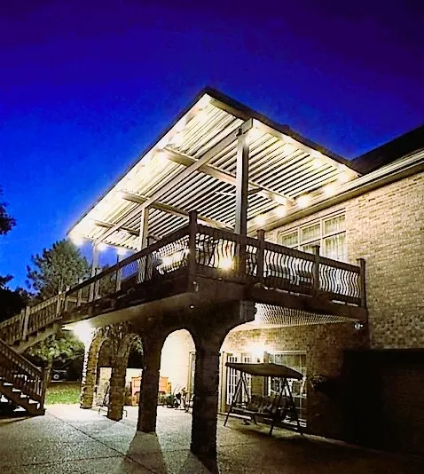 Louvered roof lit from below over an upper deck with a dark railing and arched stone supports, at night
