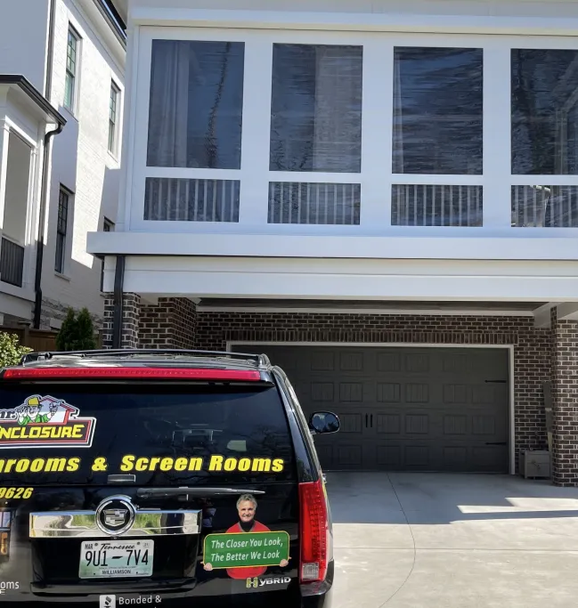 The Mr. Enclosure Sunrooms and Screen Rooms SUV parked in a driveway below a glass room built over a garage