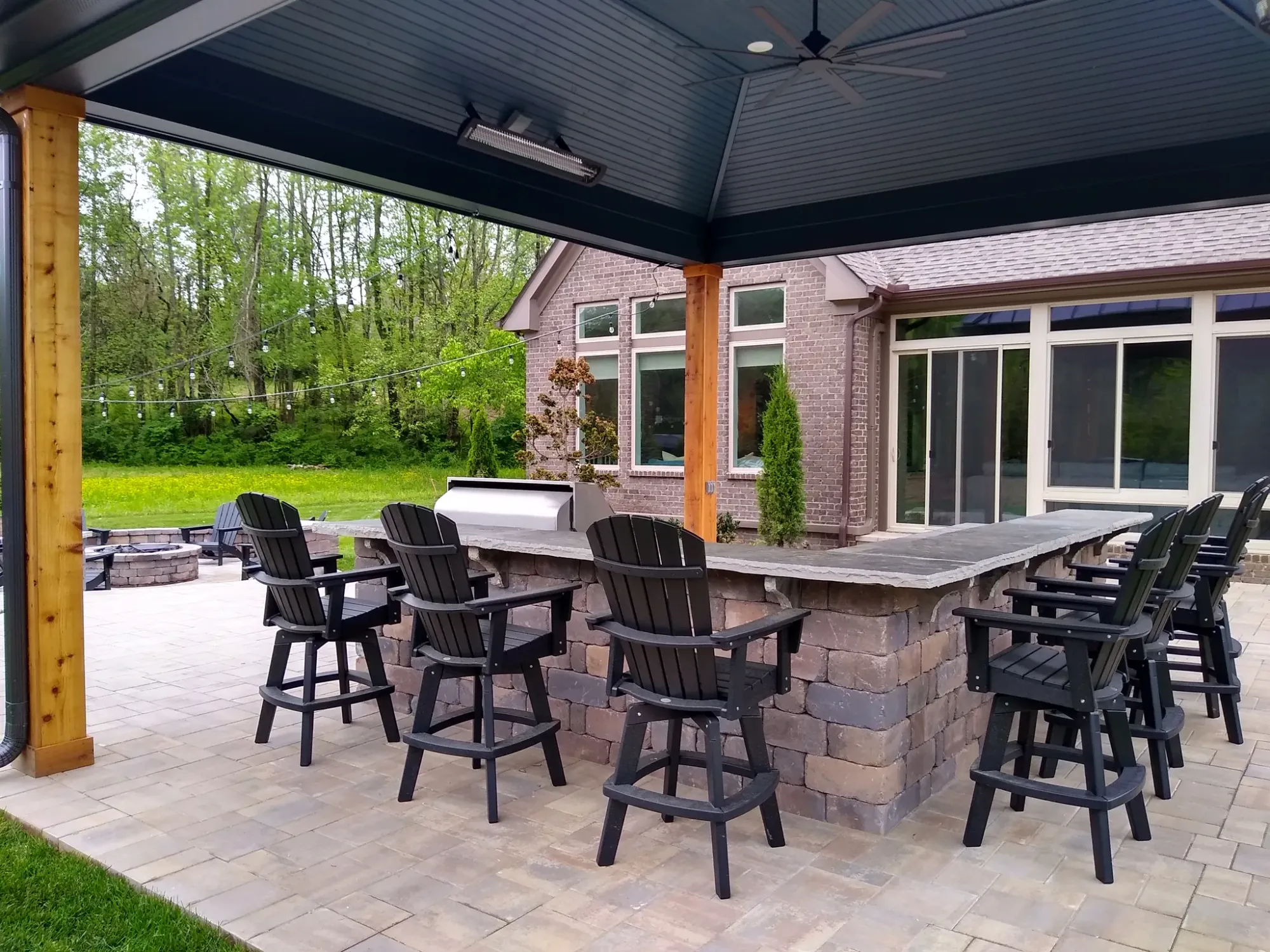 Stone outdoor kitchen bar with black swivel bar stools under a pavilion roof on stained wood posts, a brick house behind