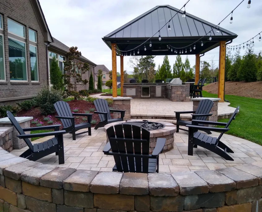 Round paver patio with Adirondack chairs around a fire pit, and a pavilion kitchen with string lights beyond it