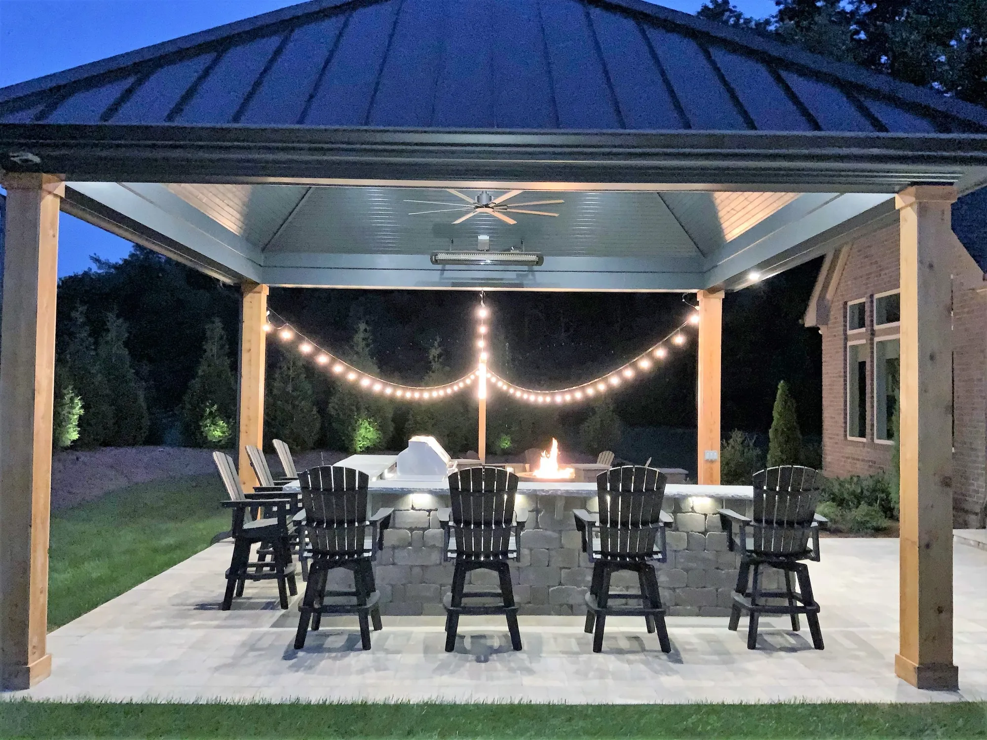 Pavilion kitchen at night with string lights, a glowing fire feature on the counter and bar chairs on the patio