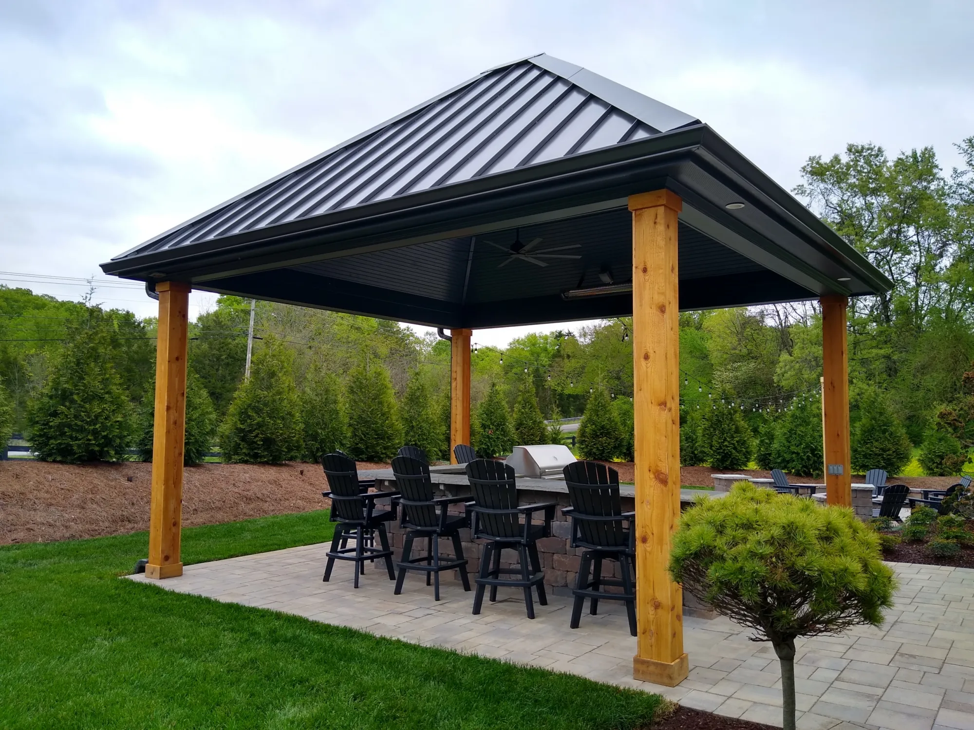 Dark standing seam pavilion roof on wood posts over a paver patio with a bar and chairs