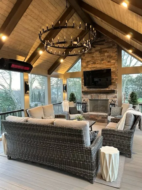 Porch with electric heaters on the beams, a round iron chandelier, wicker seating and a stone fireplace with a television