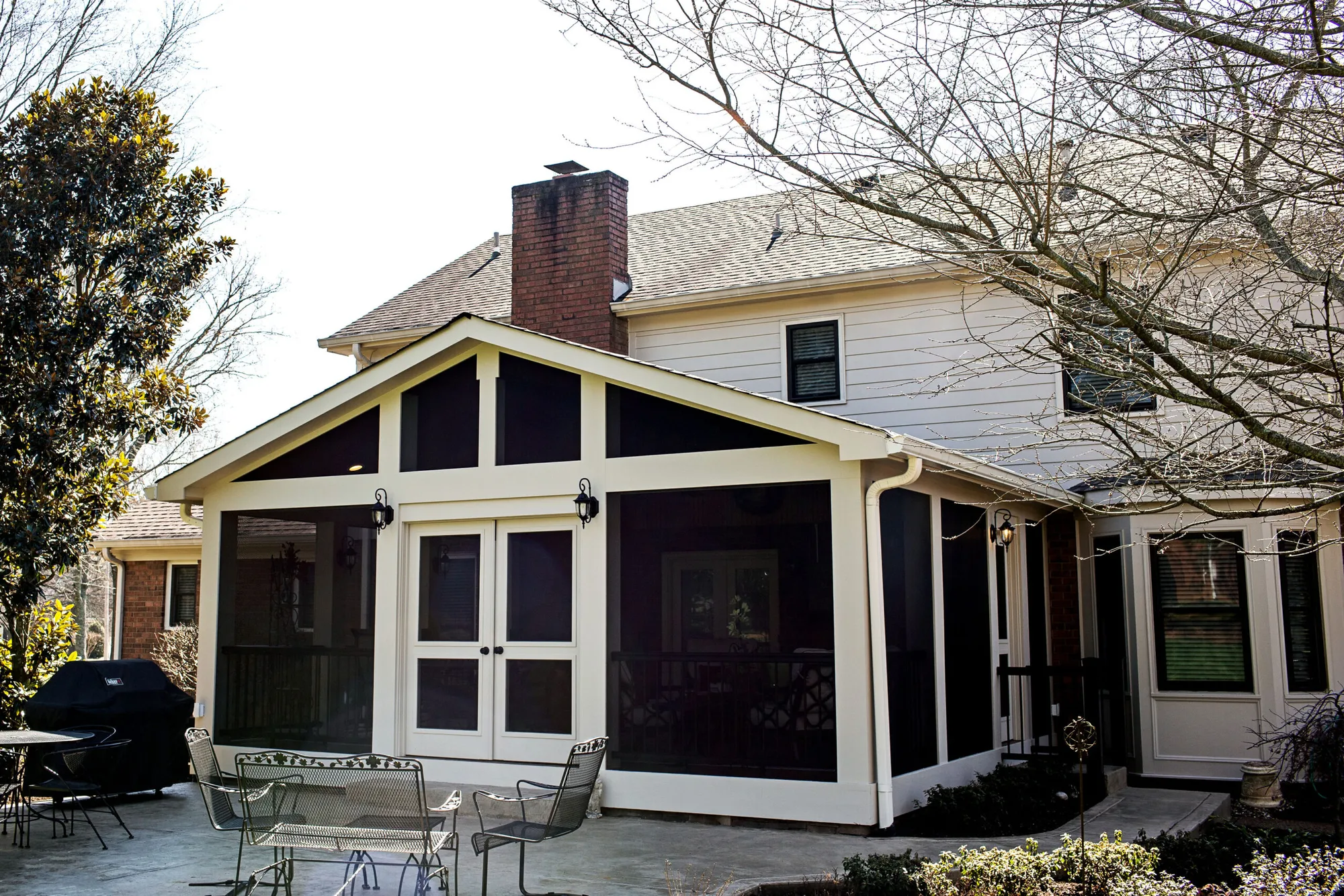 Gable screen room with white trim, double French doors and a brick chimney behind it, on a stone patio with a grill and chairs