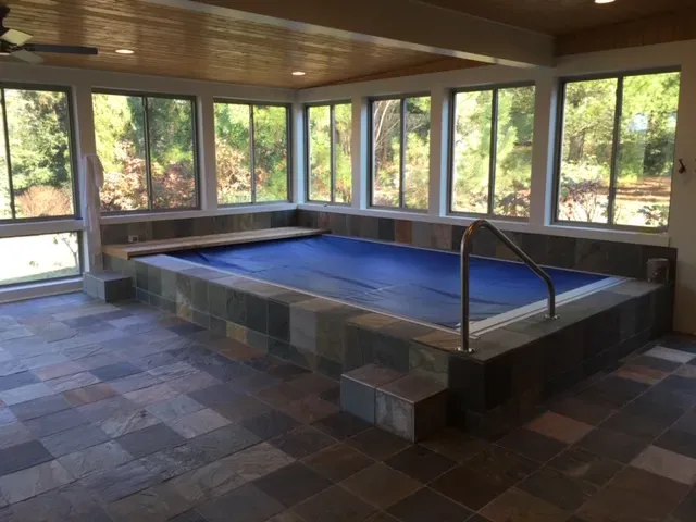 Indoor swim spa under a blue cover, set in a raised slate tile surround with steps and a grab rail, windows all around