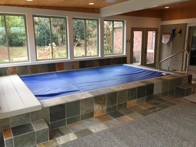 Slate tile surround of an indoor swim spa with a white bench, windows to the yard and French doors at the end