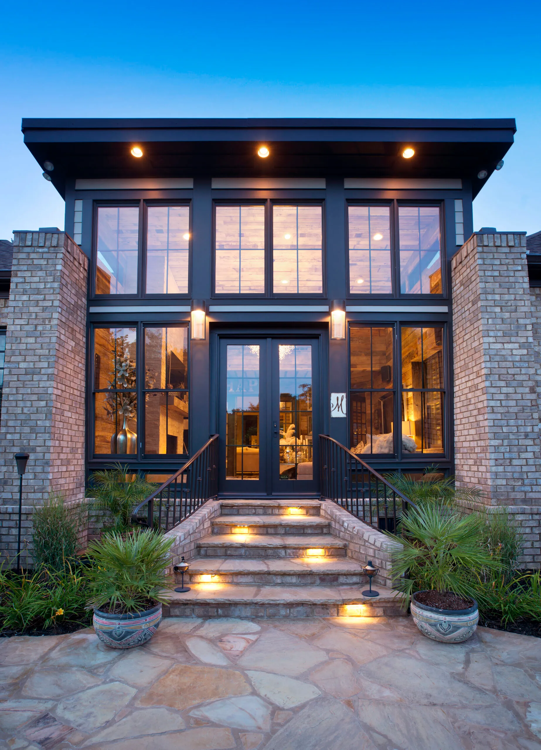 Two-story glass sunroom with black frames and French doors between brick piers, lit at dusk above flagstone steps