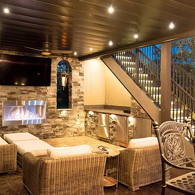 Room under a deck with a dark ceiling and recessed lights, a stone wall with a linear fireplace and television, and lit stairs